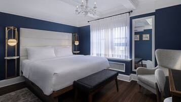Una camera d'albergo con un letto grande, una panca, una sedia, un lampadario e un'ampia finestra con tende leggere.