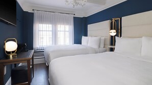 Habitación de hotel con dos camas, un candelabro, un escritorio con lámpara y un ventanal grande con cortinas.