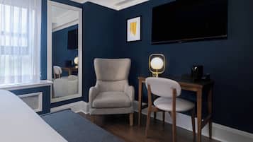 Habitación de hotel con una cama grande, un sillón gris, un escritorio de madera con una lámpara, un televisor de pantalla plana y una obra de arte enmarcada en la pared.