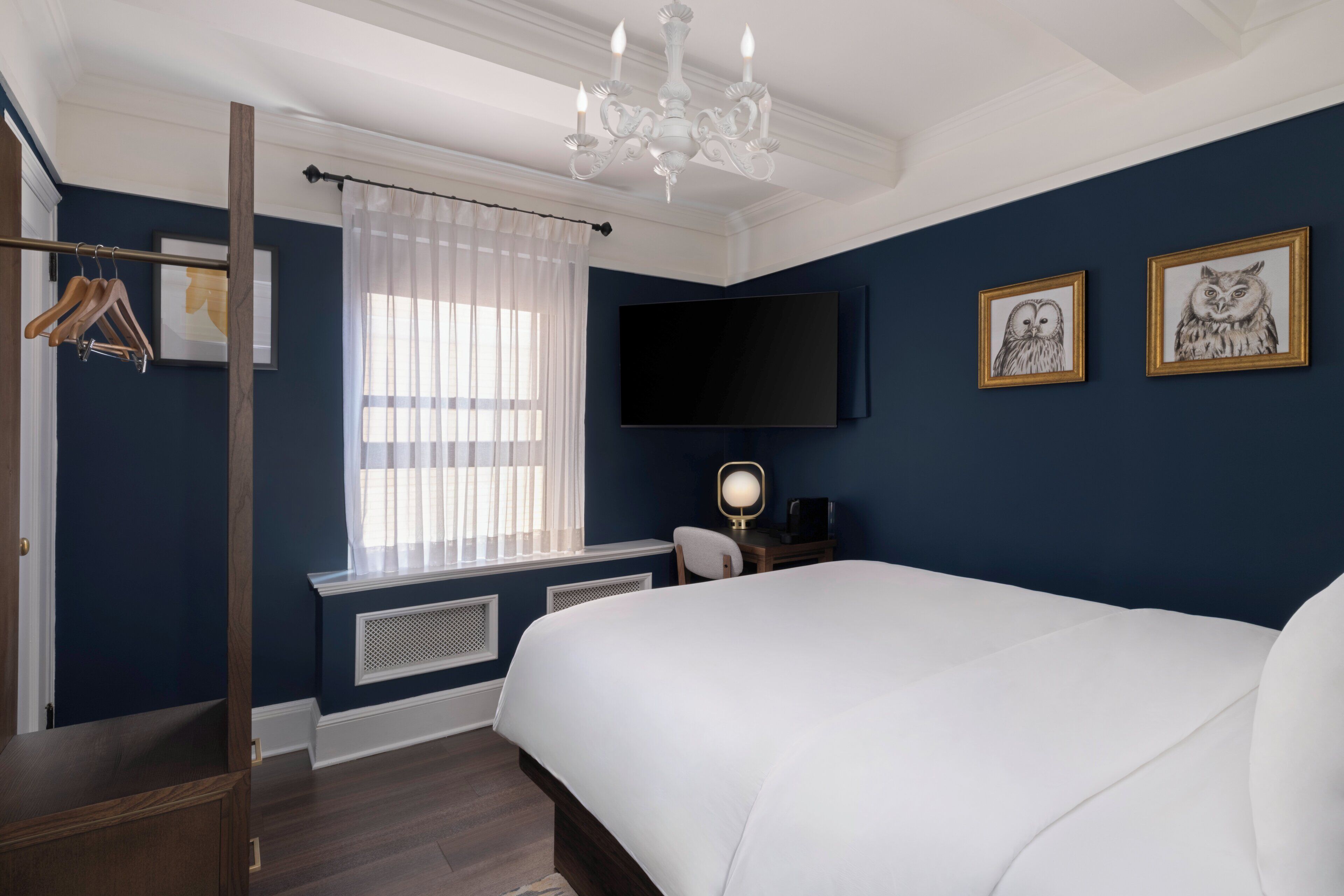 Un dormitorio con una cama grande, un televisor montado en la pared, un escritorio con una silla y una ventana con cortinas ligeras.