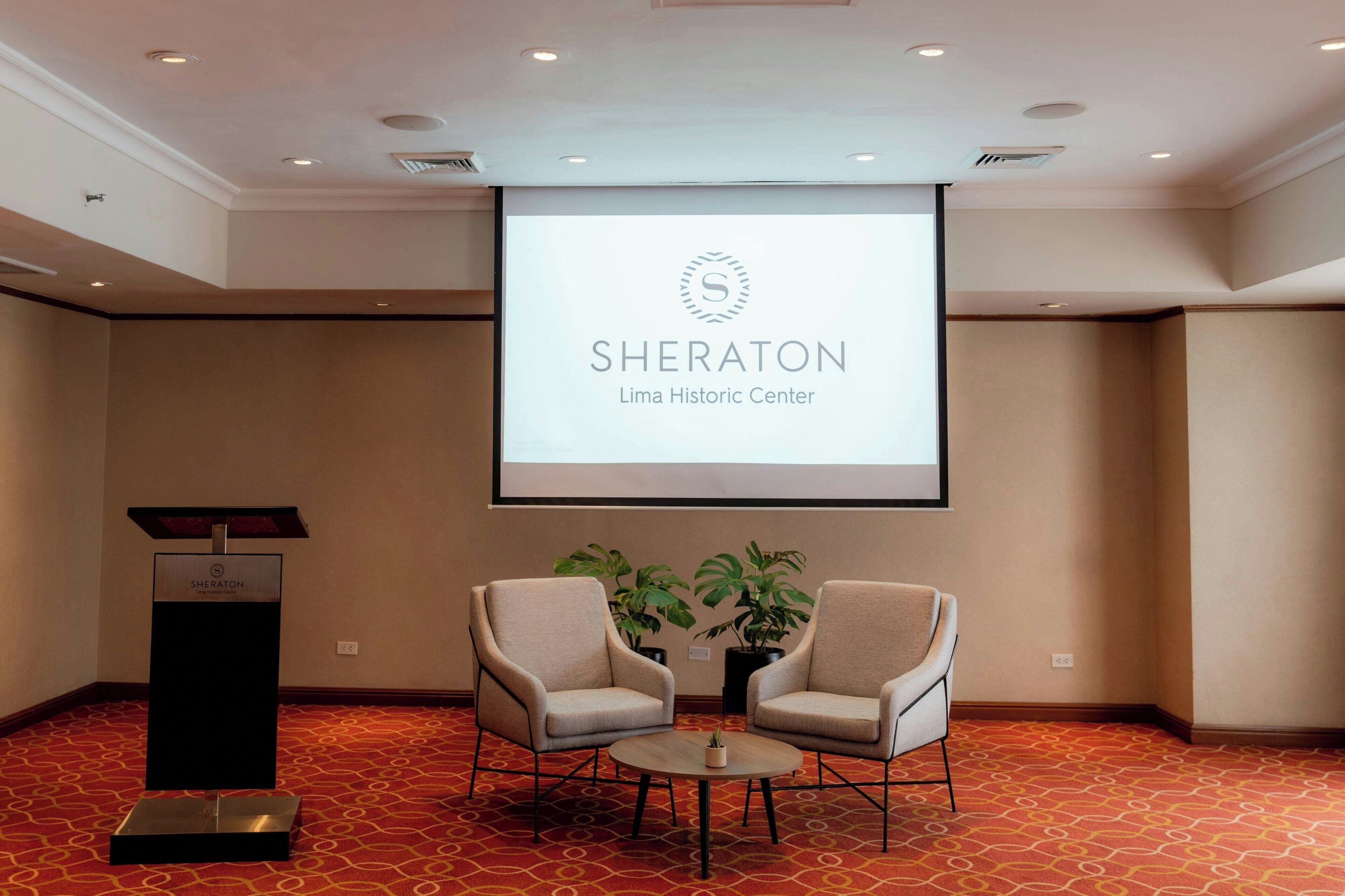 Una sala de conferencias con el logotipo de Sheraton, dos sillas, un atril y una pantalla de proyector.
