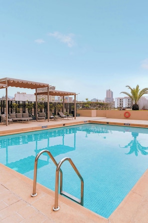Ein Pool auf dem Dach mit Liegestühlen und einer Pergola, umgeben von einer Stadtansicht.