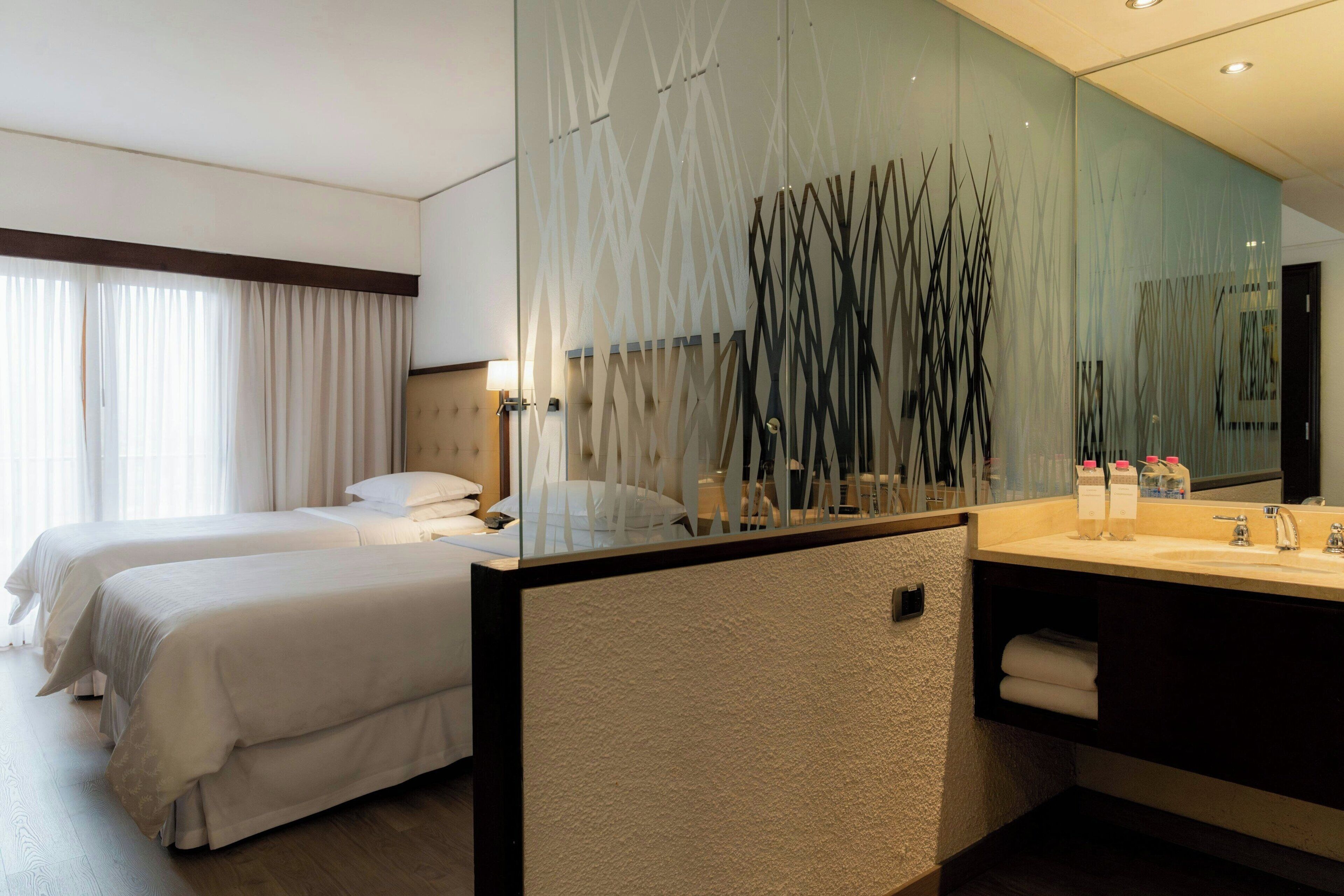 A hotel room with two beds, a large mirror, a modern bathroom with a sink and towel rack.