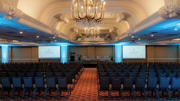 Un salón de conferencias con filas de sillas, un escenario con un atril y dos pantallas mostrando el logotipo de Sheraton.