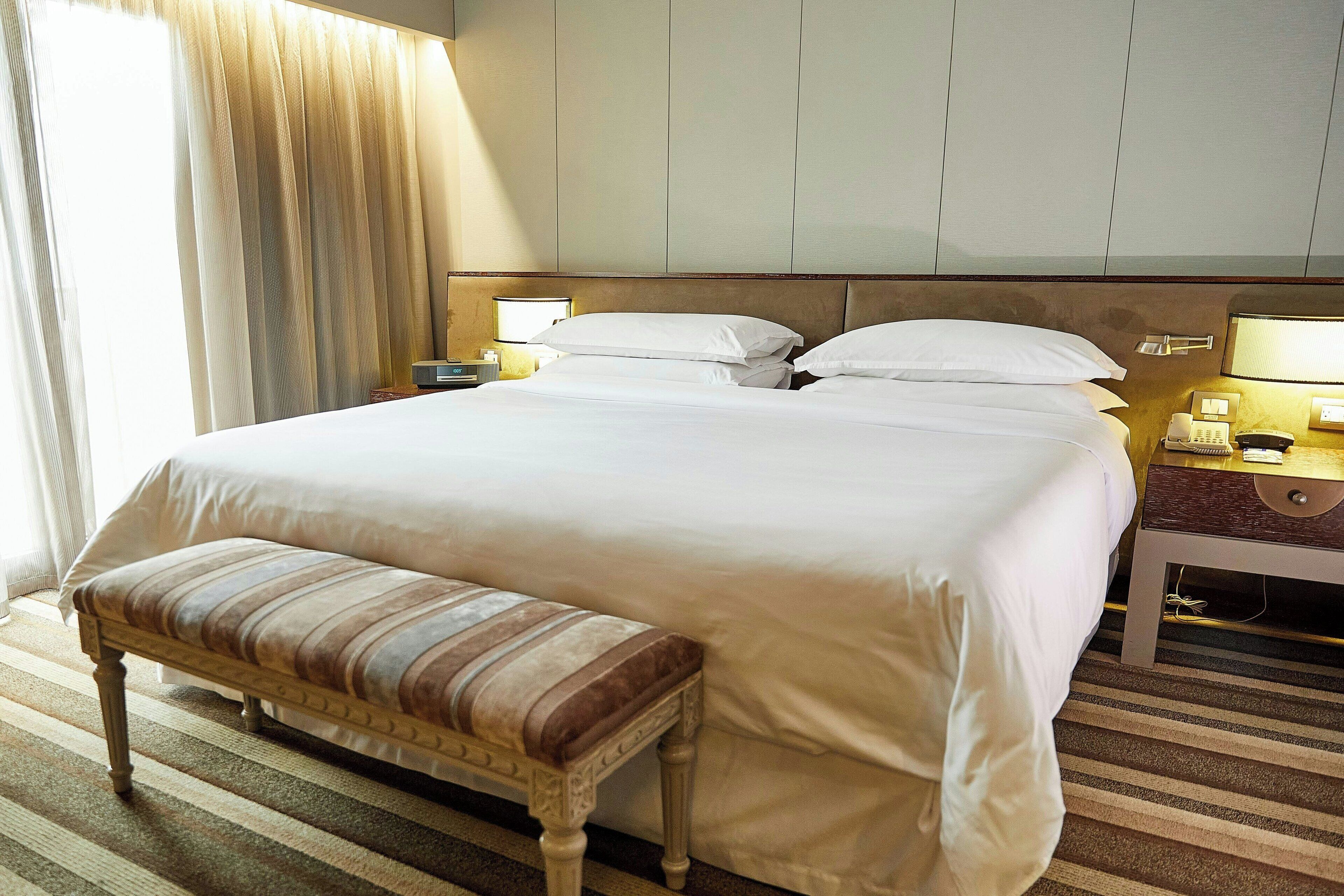 Une chambre d’hôtel avec un grand lit, une table de chevet avec une lampe, un banc et une fenêtre avec des rideaux.