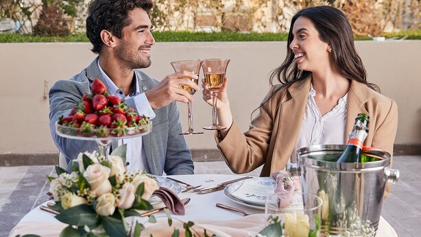 Un couple trinque au champagne à une table en plein air, agrémentée d’un arrangement floral, avec des palmiers et un bâtiment en arrière-plan.