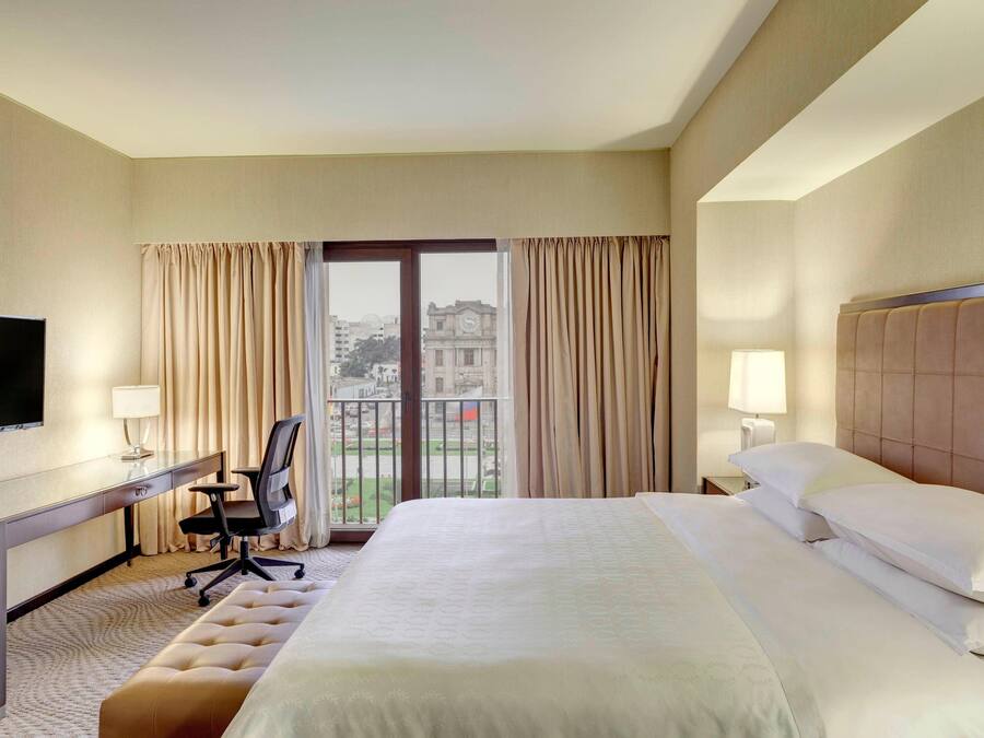 A hotel room with a large bed, a desk with a chair, a television, and a view of the city through a balcony.