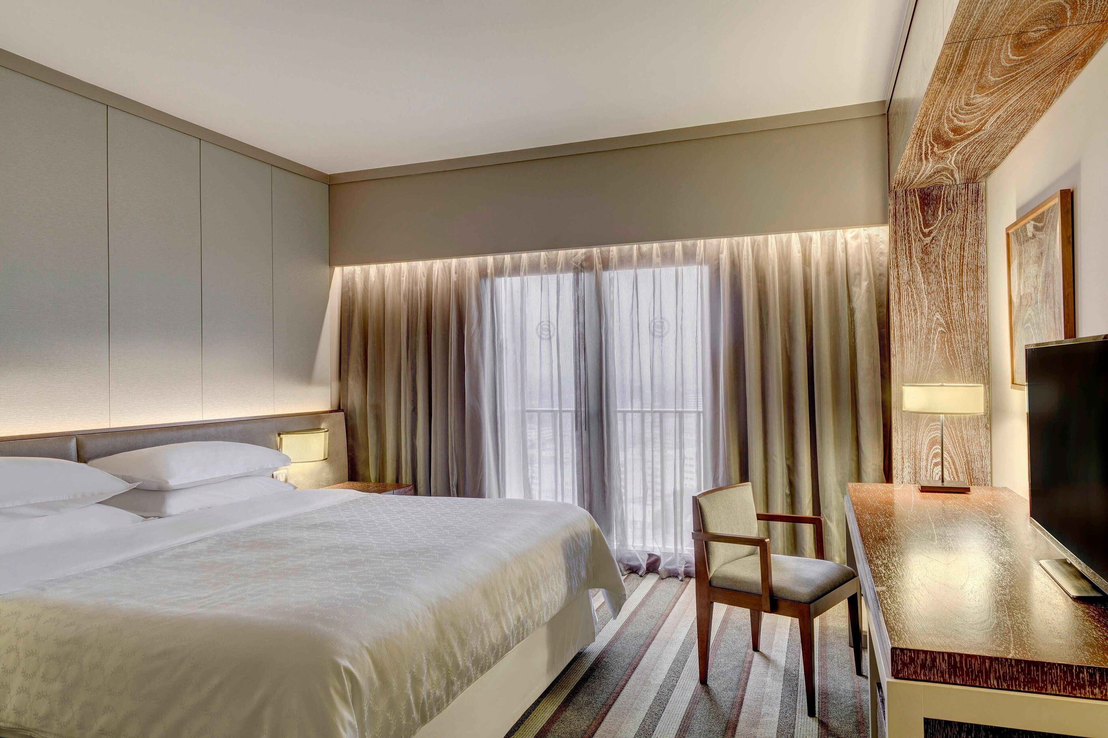 Habitación de hotel con una cama grande, un escritorio con silla, un televisor y una ventana con cortinas.
