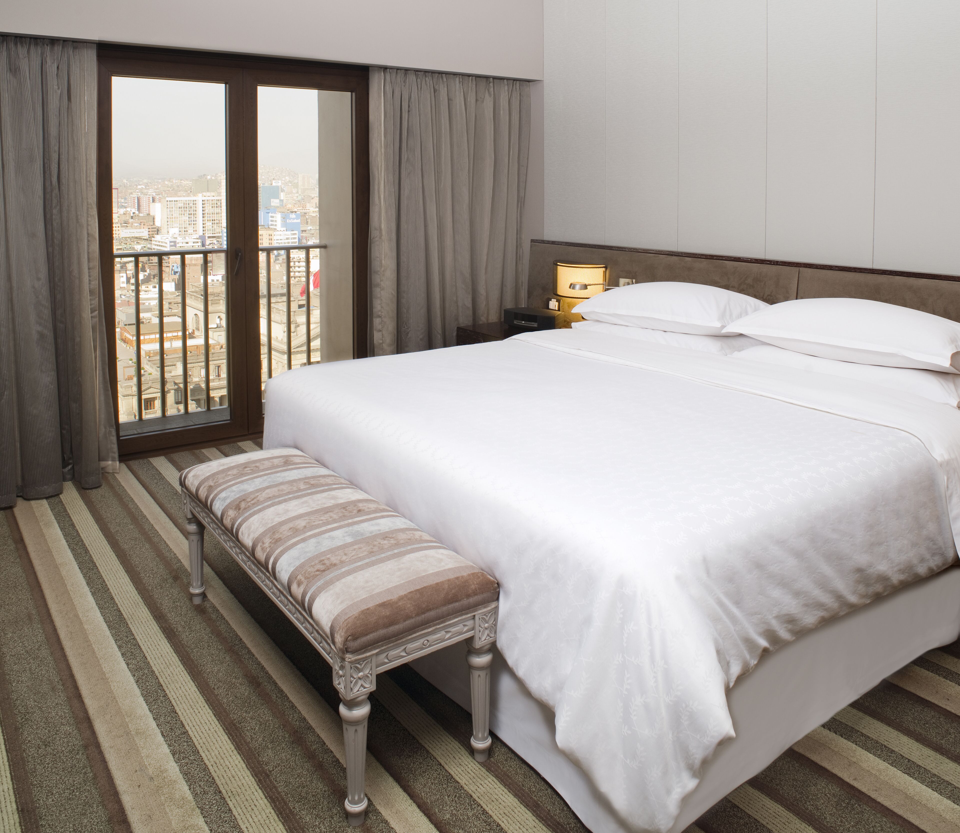 Une chambre d’hôtel avec un grand lit, une banquette, une table de chevet avec une lampe et une vue sur la ville par la fenêtre.
