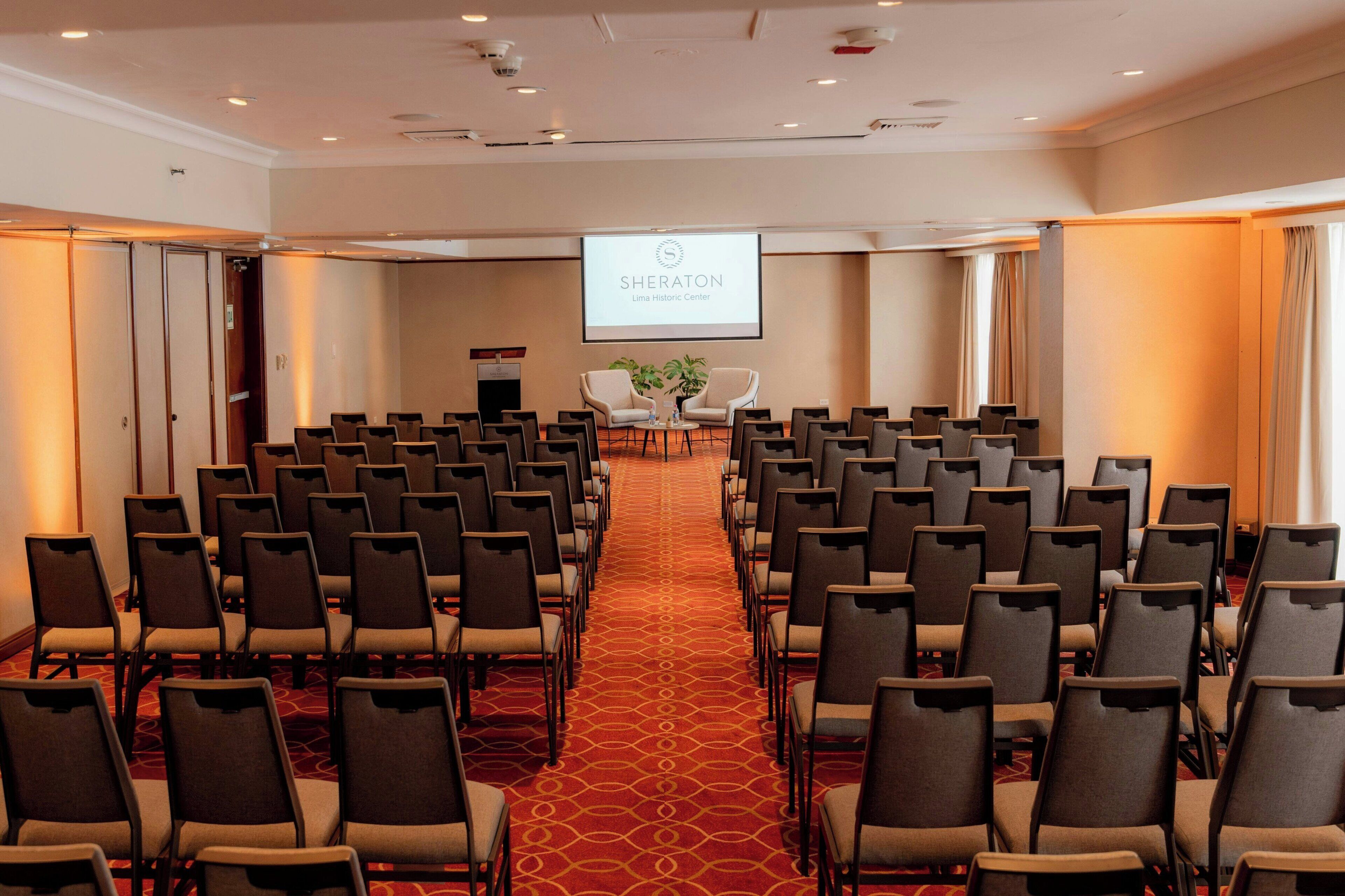 Un salón de conferencias con filas de sillas, piso alfombrado, un atril y una pantalla de proyección mostrando 'SHERATON Lima Historic Center'.