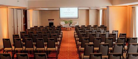 Un salón de conferencias con filas de sillas, piso alfombrado, un atril y una pantalla de proyección mostrando 'SHERATON Lima Historic Center'.