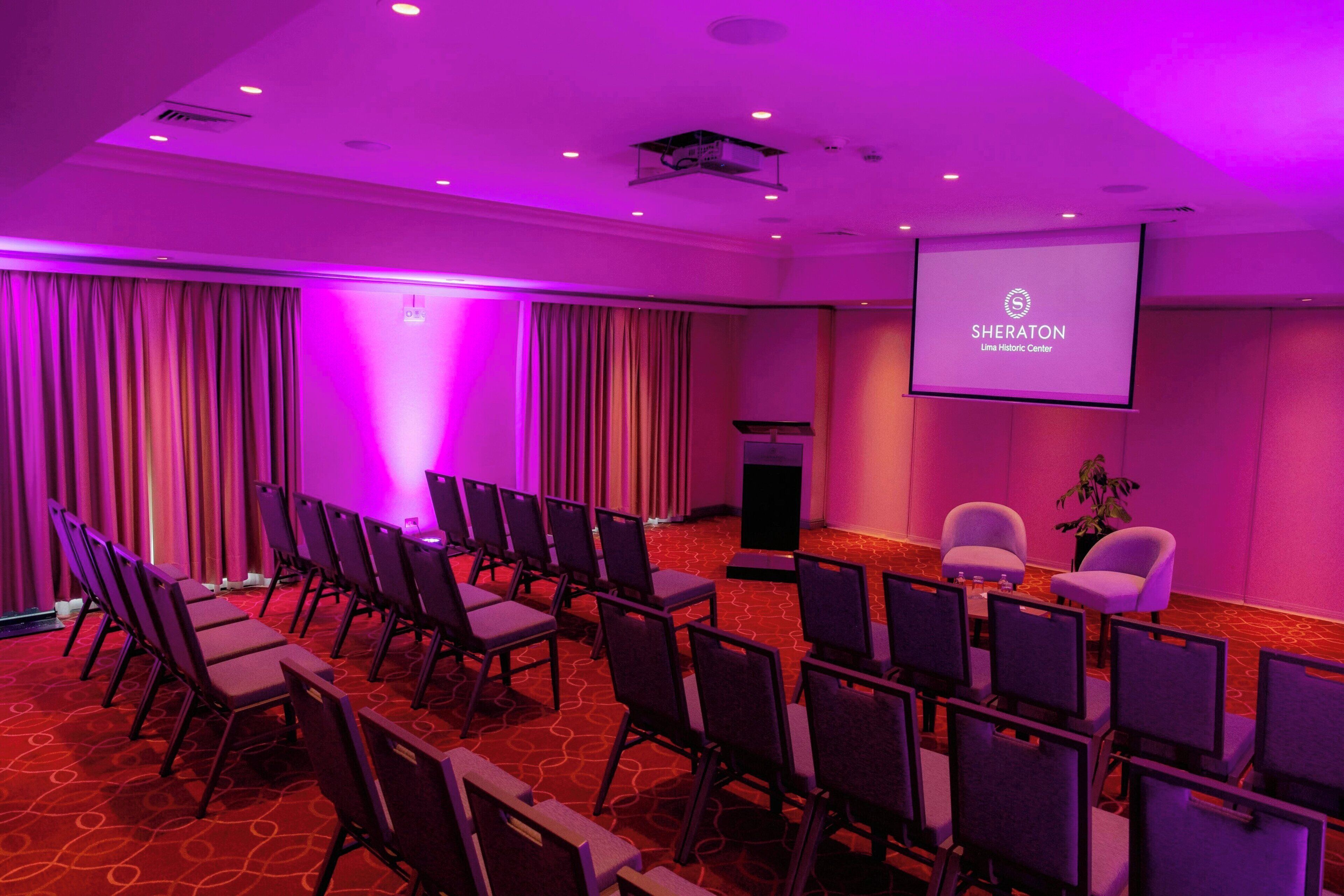Une salle de conférence avec des rangées de chaises, un écran de projection et un espace dédié aux présentations.