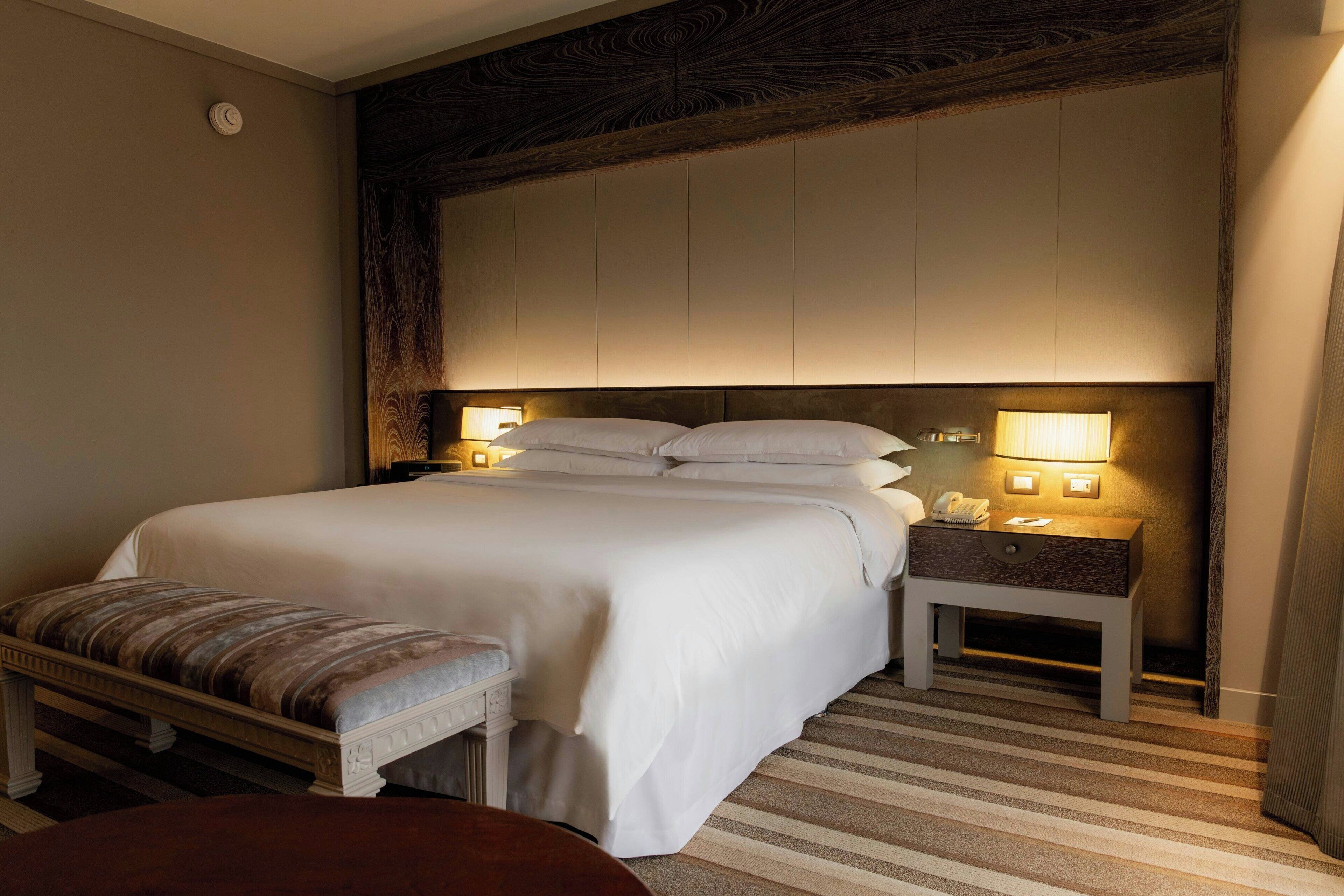 A hotel room with a large bed, bedside lamps, a bench, and a nightstand.