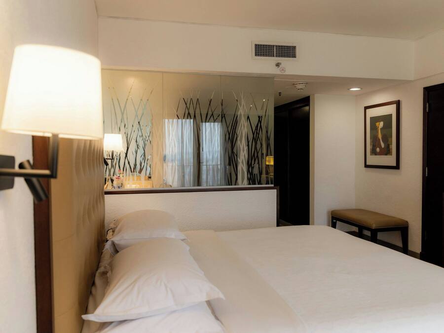 Quarto de hotel com cama, um banquinho, um espelho e um quadro na parede.