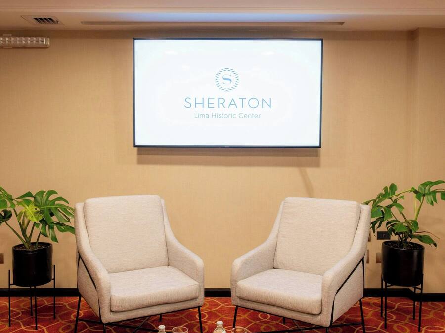 Un salón de conferencias con dos sillones, una mesita con botellas de agua y una pantalla de proyector mostrando 'SHERATON Lima Historic Center'.
