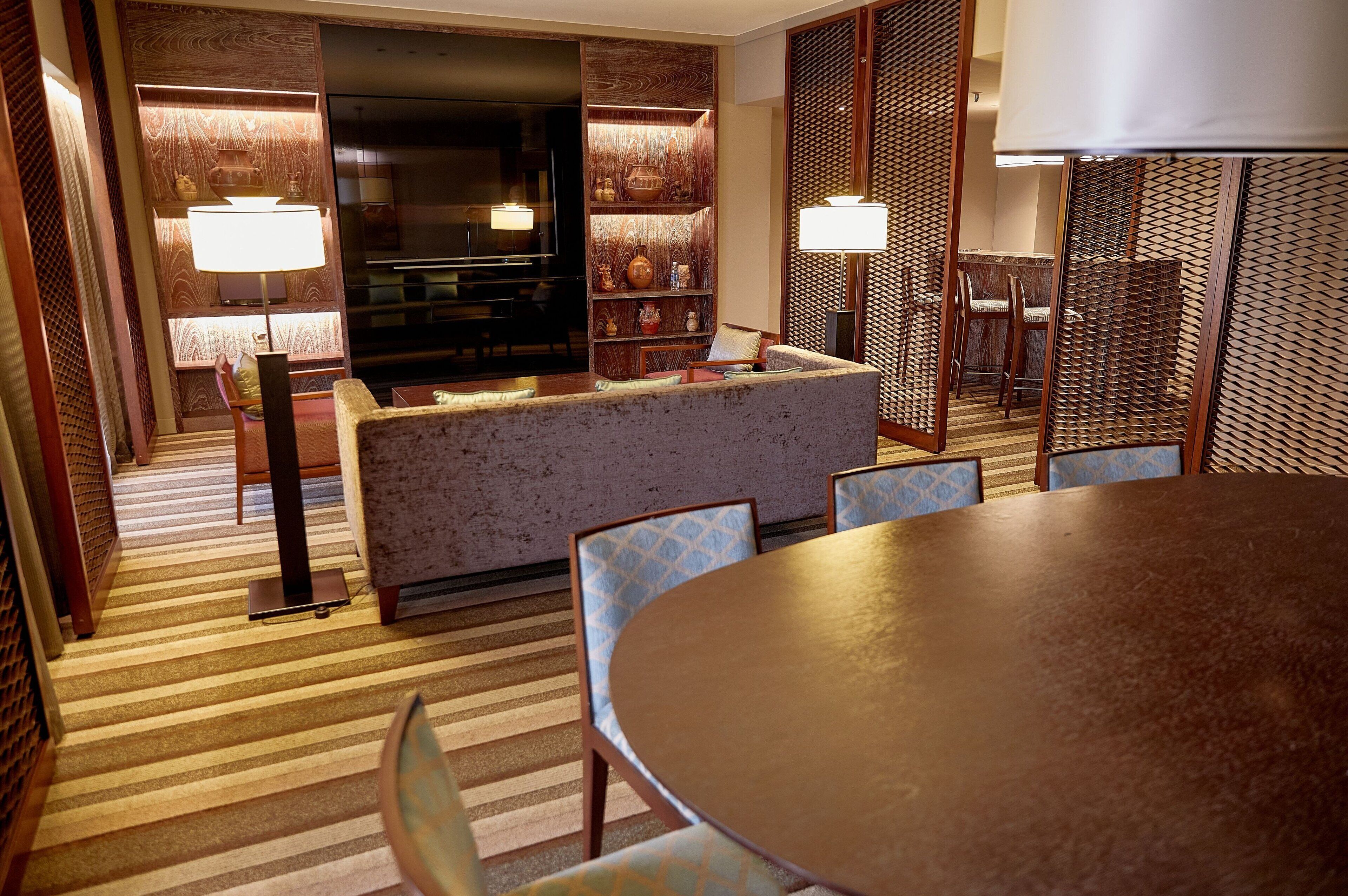 A hotel room with a sofa, dining table, and chairs, a TV, and a bookshelf with decorative items.