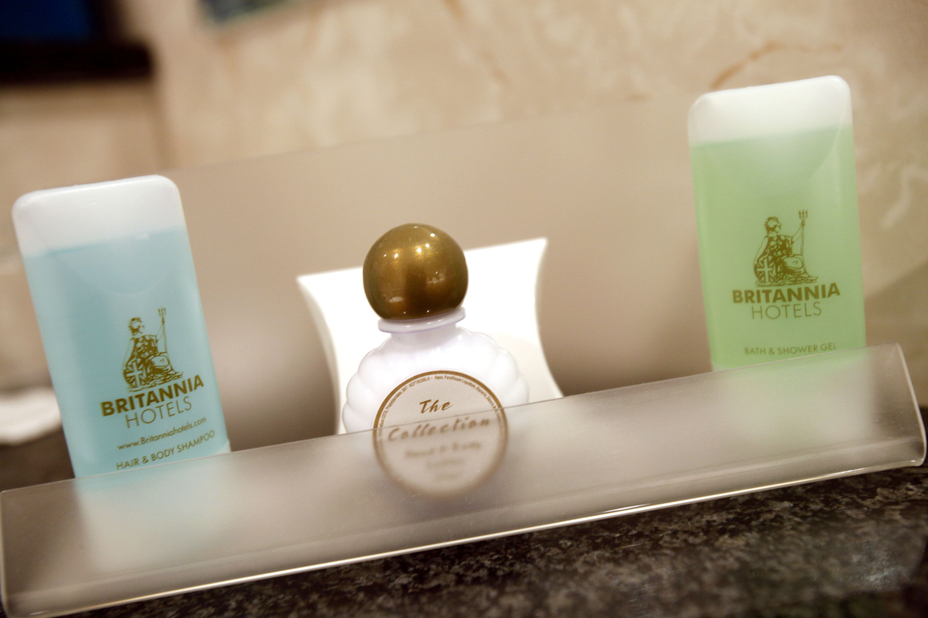Three toiletries on a hotel counter: a blue bottle labeled 'Britannia Hotels Hair & Body Shampoo', a clear bottle with a gold cap labeled 'The Collection', and a green bottle labeled 'Britannia Hotels Bath & Shower Gel'.