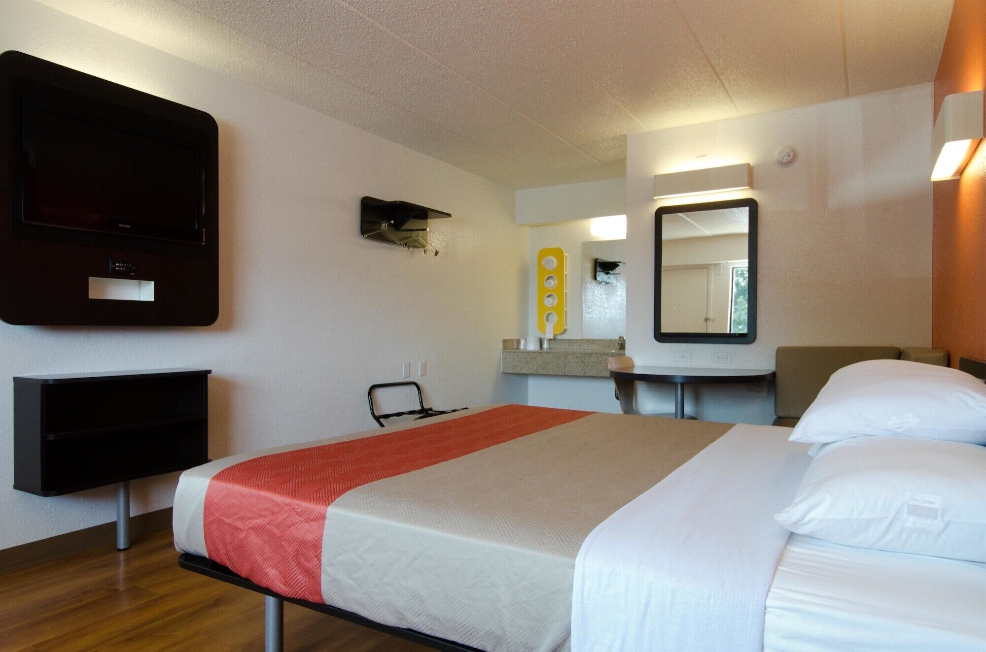 A hotel room with a bed, a TV, a mirror, a small desk, and a wall-mounted shelf.