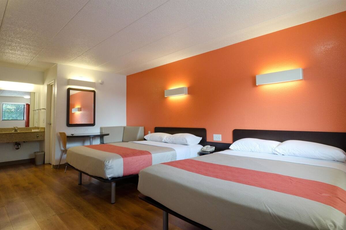 A hotel room with two beds, a small table, a chair, a mirror, and a window.