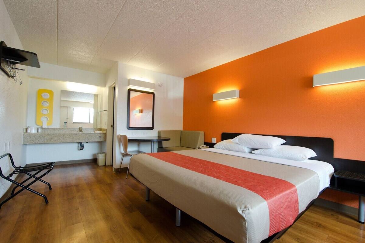 A hotel room with a bed, a desk, a chair, a small table, a mirror, and a bathroom with a sink and toilet.