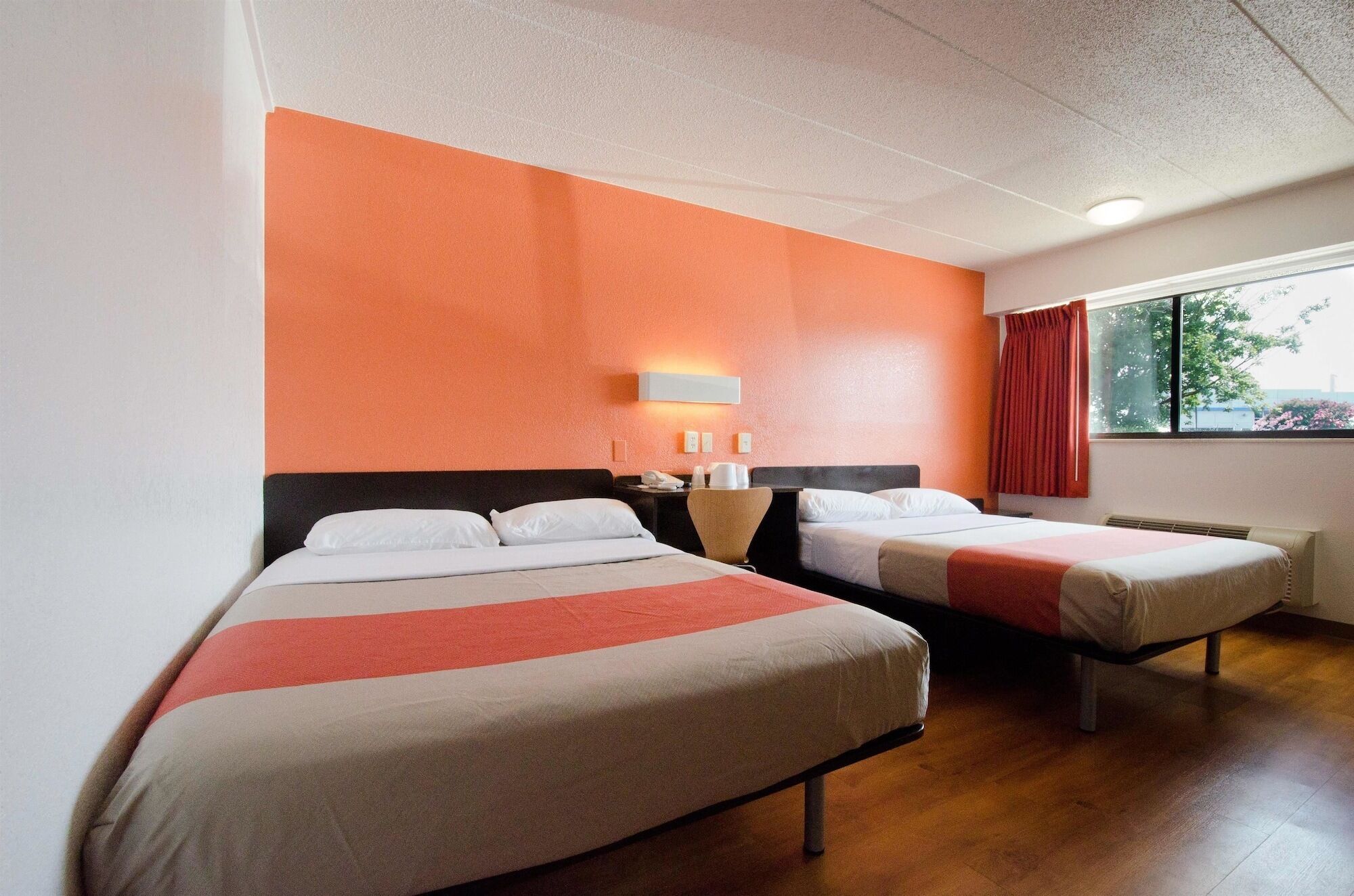Quarto de hotel com duas camas, mesa de cabeceira, luminária e janela com cortinas.