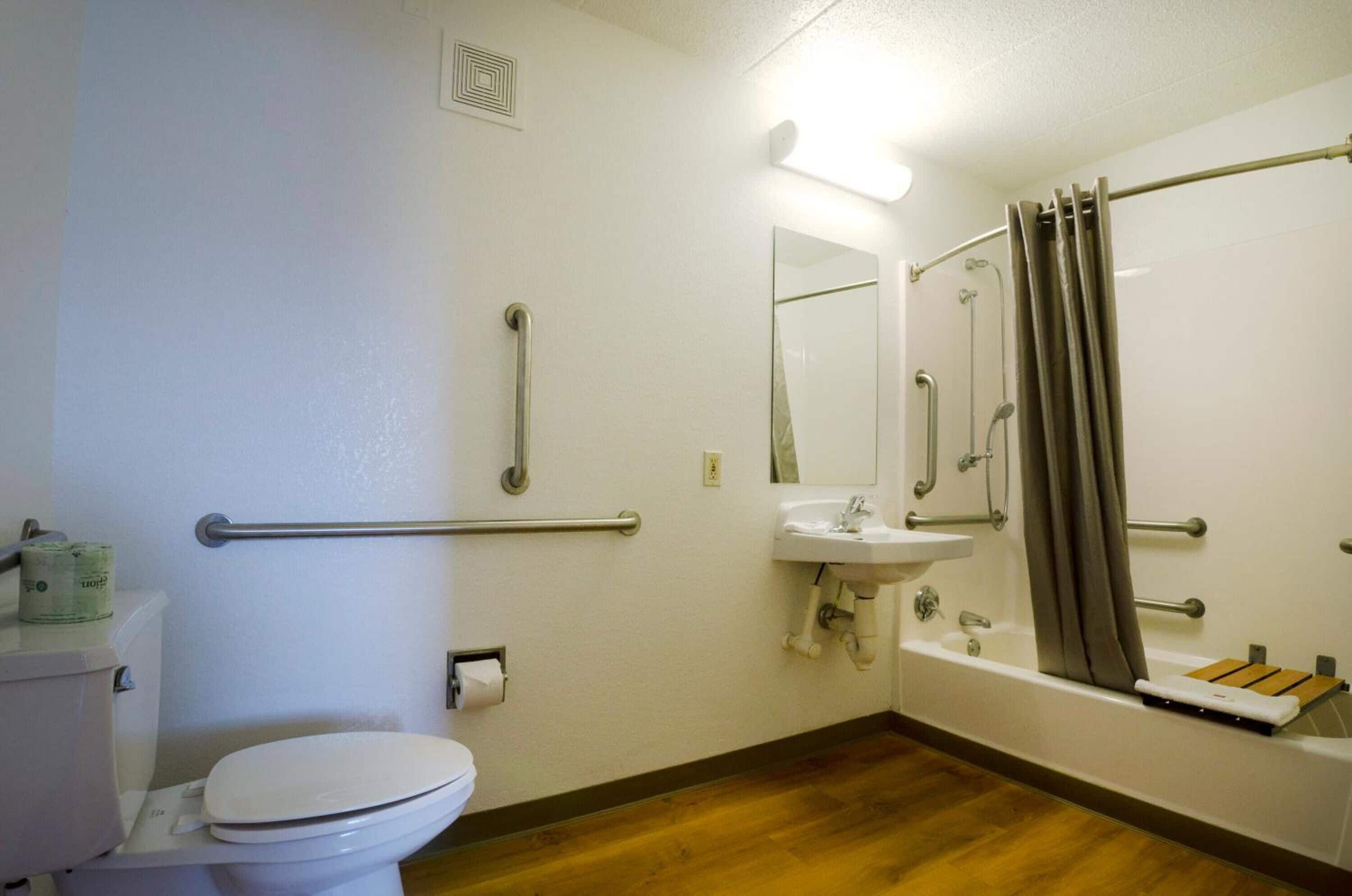 Ein Badezimmer mit Toilette, Waschbecken und Dusche. Es sind Haltegriffe und ein Handlauf vorhanden.