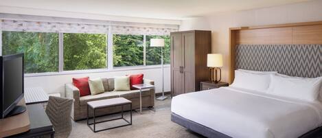 A hotel room with a bed, a desk with a computer, a sofa, and a window with a view of trees.