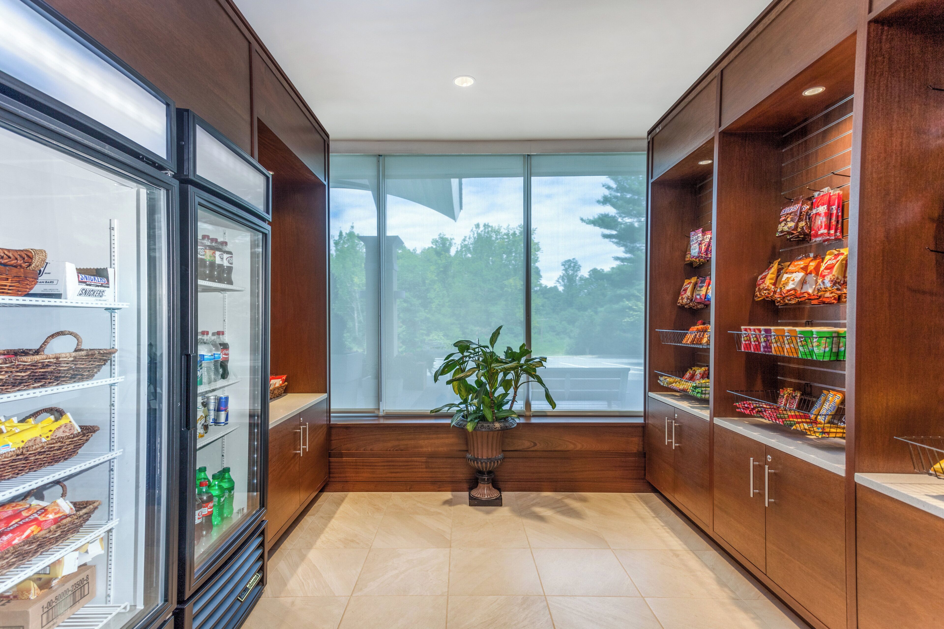 A store interior with shelves stocked with snacks and beverages, a refrigerated display case, and a potted plant in the center.