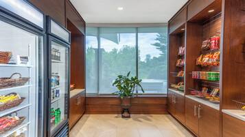 A store interior with shelves stocked with snacks and beverages, a refrigerated display case, and a potted plant in the center.