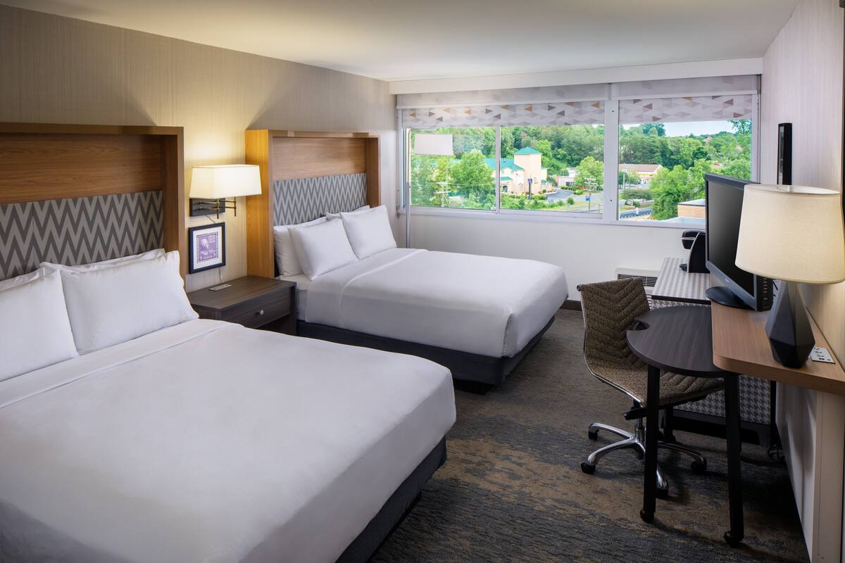 A hotel room with two beds, a desk with a computer, a TV, and a window with a view of the city.