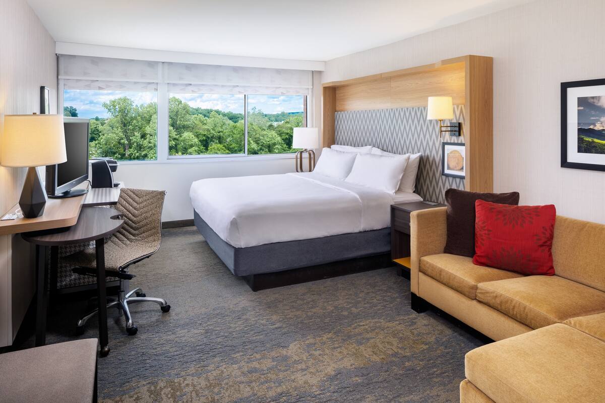 A hotel room with a large bed, a desk with a computer, a chair, a sofa, and a view of greenery through the window.
