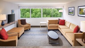 A modern living room with a sofa, armchairs, a coffee table, and a TV. There are large windows with a view of trees. - Holiday Inn Charlottesville-Monticello by IHG (Charlottesville)