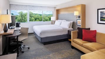 Une chambre d’hôtel avec un lit, un bureau avec un ordinateur, une chaise, un canapé et une fenêtre donnant sur des arbres.