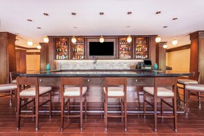 A bar area with a long counter, multiple stools, a television screen, and shelves stocked with bottles. - Holiday Inn Charlottesville-Monticello by IHG (Charlottesville)