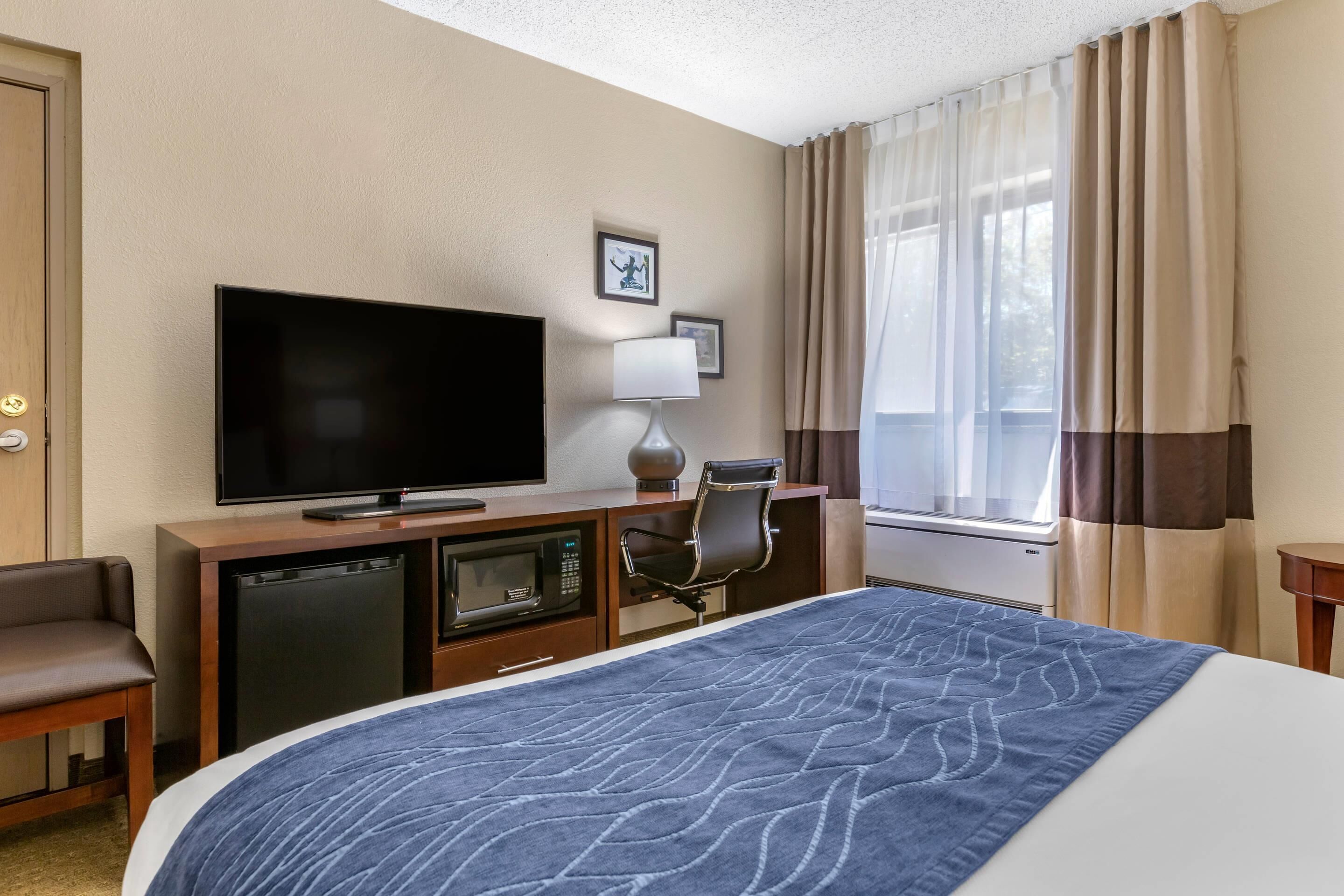 A hotel room with a bed, a television, a desk with a lamp, a chair, a microwave, and a window with curtains.