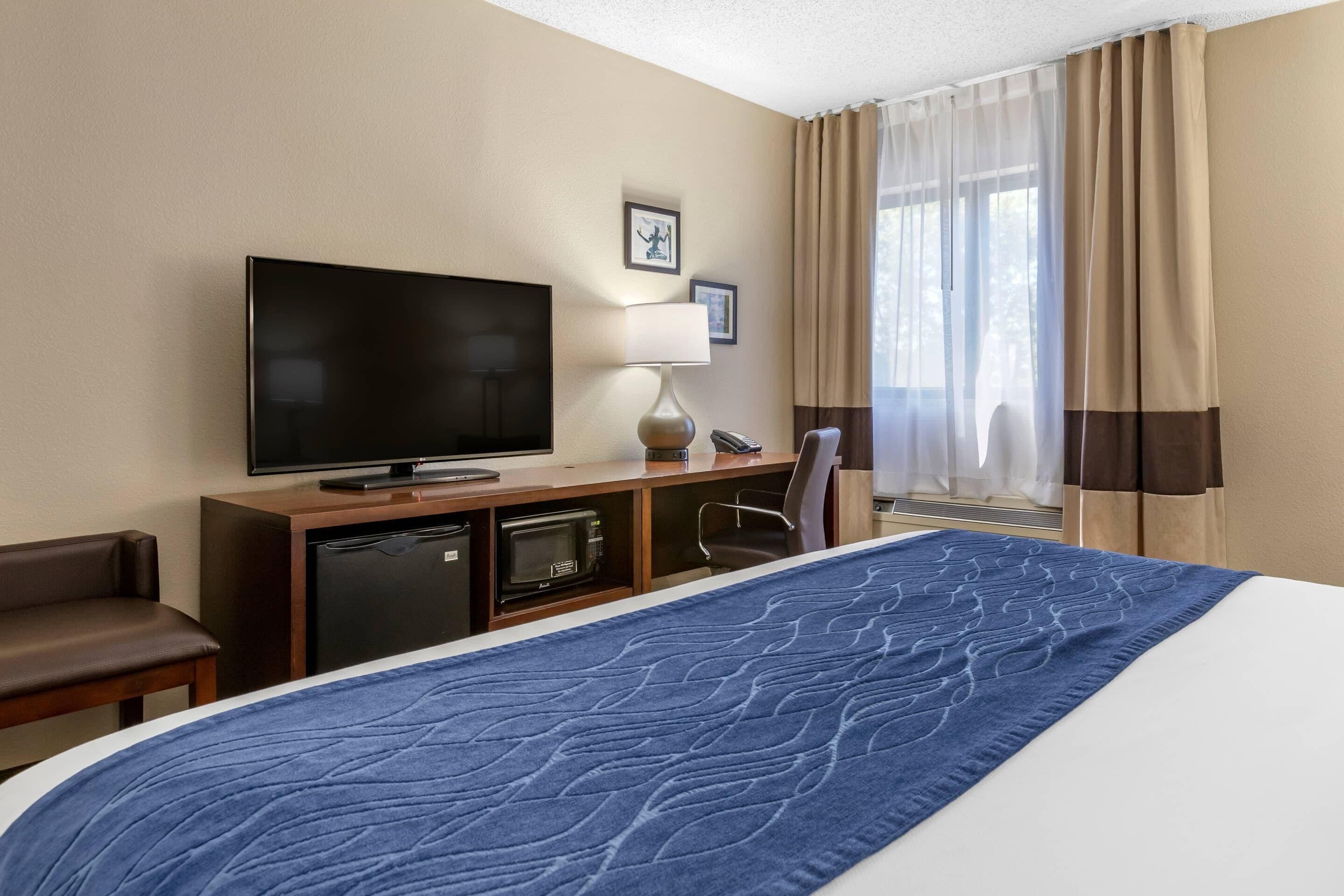 A hotel room with a bed, a television, a desk with a lamp, a chair, and a window with curtains.