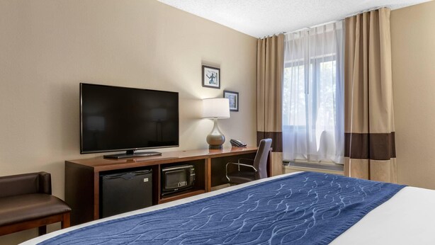 A hotel room with a bed, a television, a desk with a lamp, a chair, and a window with curtains. - Comfort Inn Near Greenfield Village (Dearborn)