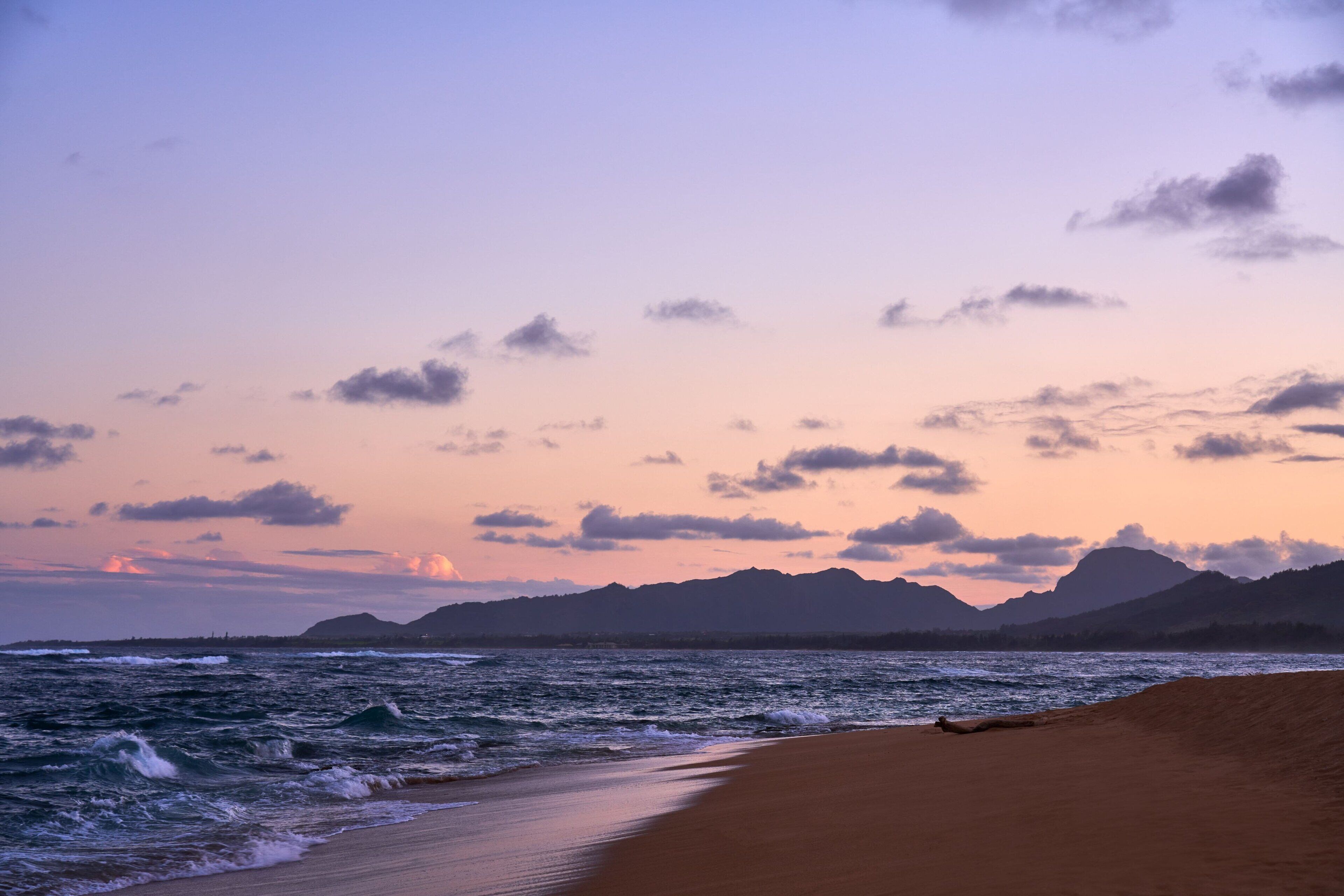 A beach with waves, a clear sky, and mountains in the background.
