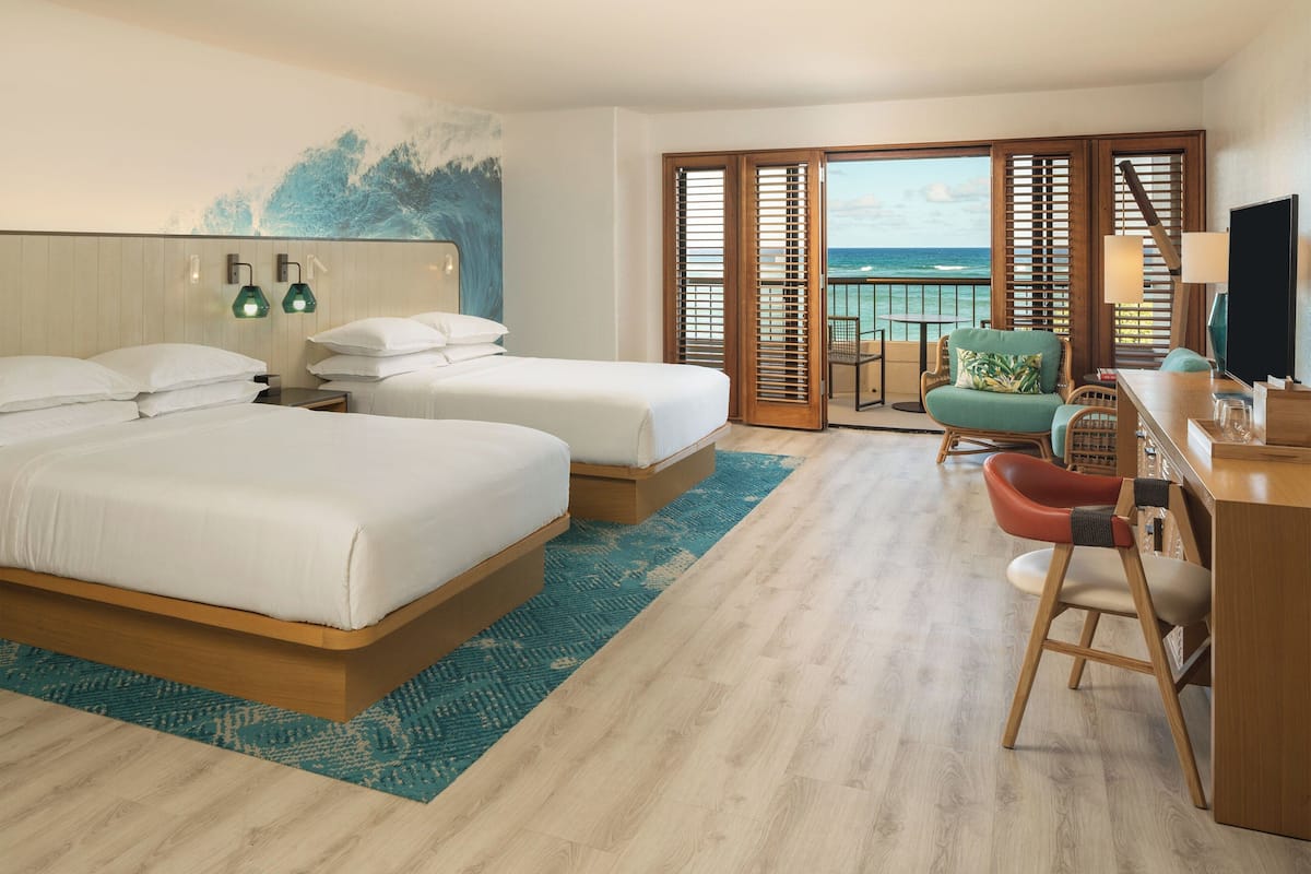 A hotel room with two beds, a desk with a chair, a TV, and a balcony with a view of the ocean.