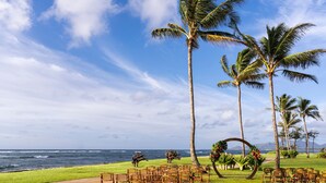 An outdoor wedding setup with chairs arranged in rows facing a decorated arch, palm trees, and a view of the ocean. - Sheraton Kauai Coconut Beach Resort (Kapaa)