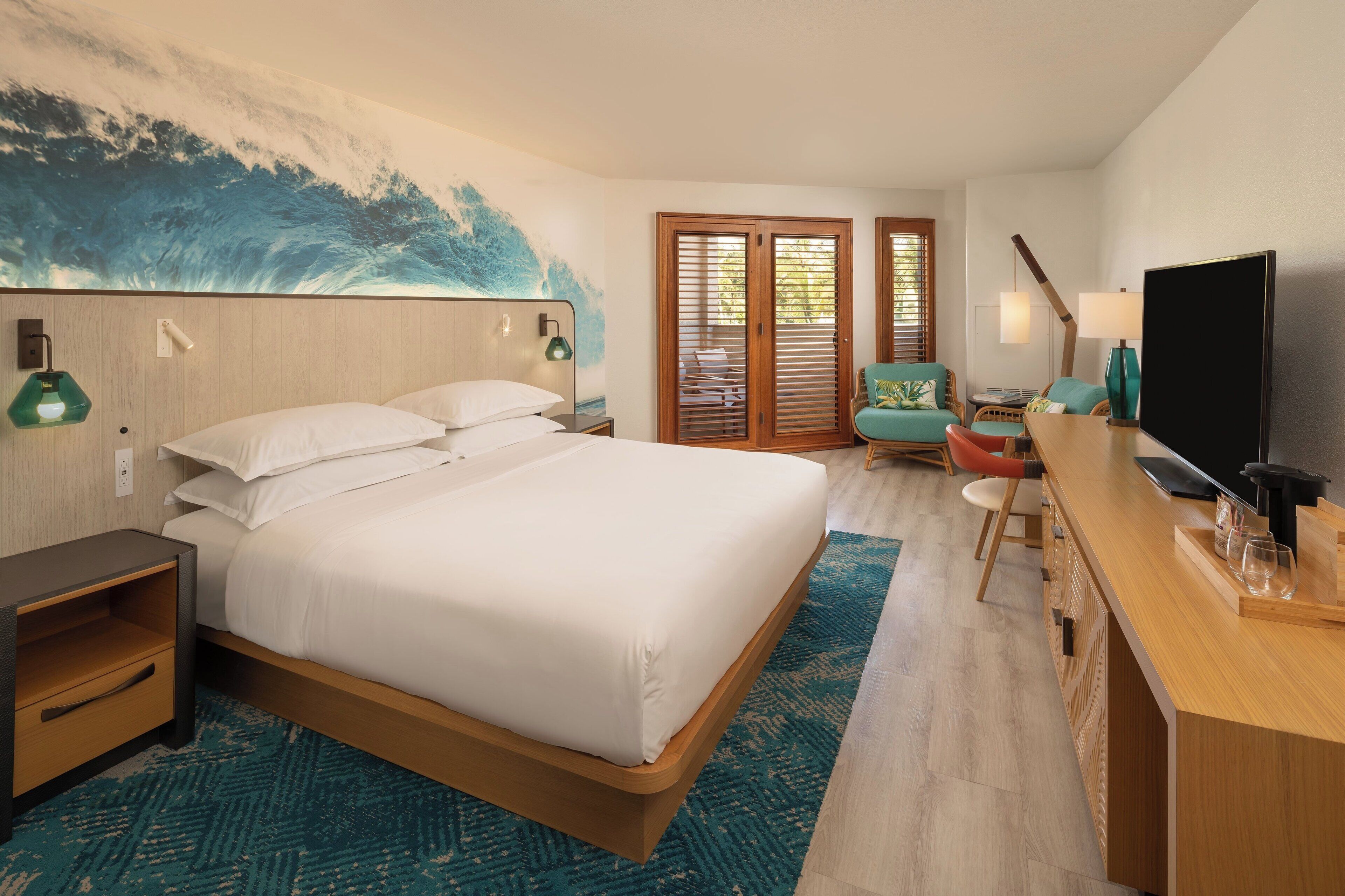 A hotel room with a large bed, a desk with a flat-screen TV, a chair, and a window with blinds.
