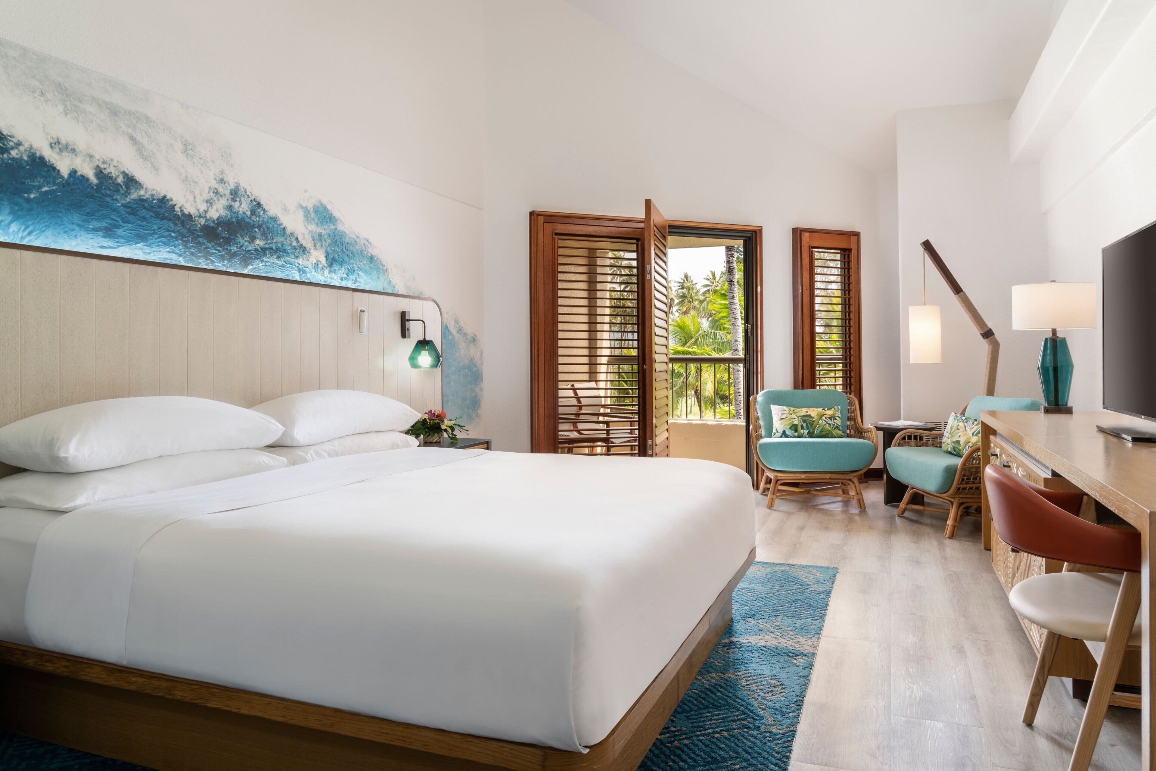 A hotel room with a large bed, a desk with a chair, a TV, and a window with a view of palm trees.