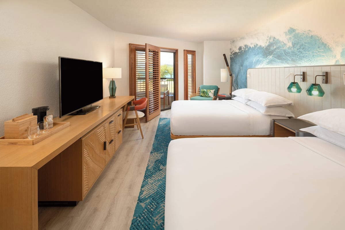 A hotel room with a TV, wooden desk, two beds, a desk chair, a small table, and a view of the outdoors.