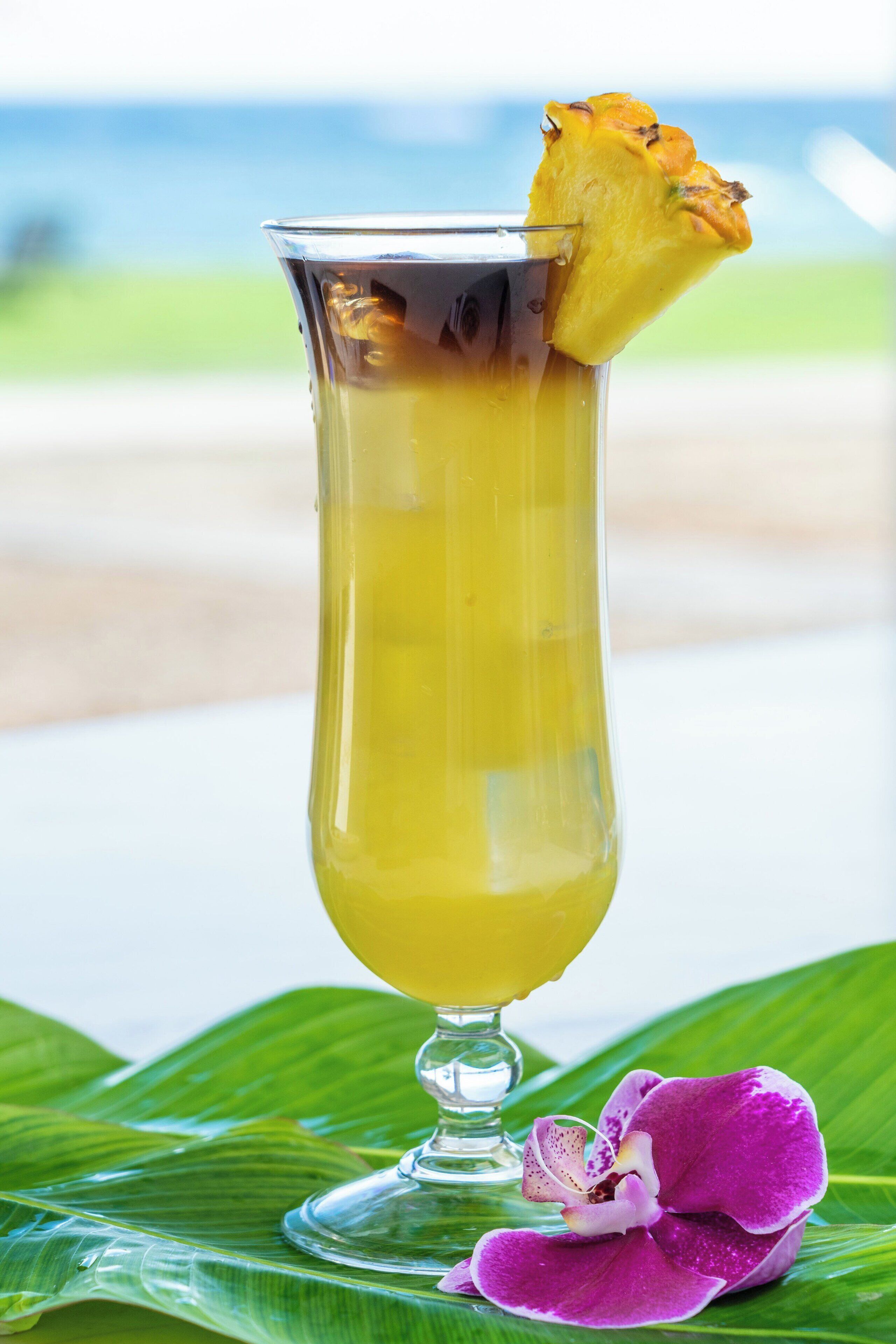 A tall glass with a yellow beverage, a pineapple slice, and a purple orchid on a green leaf.