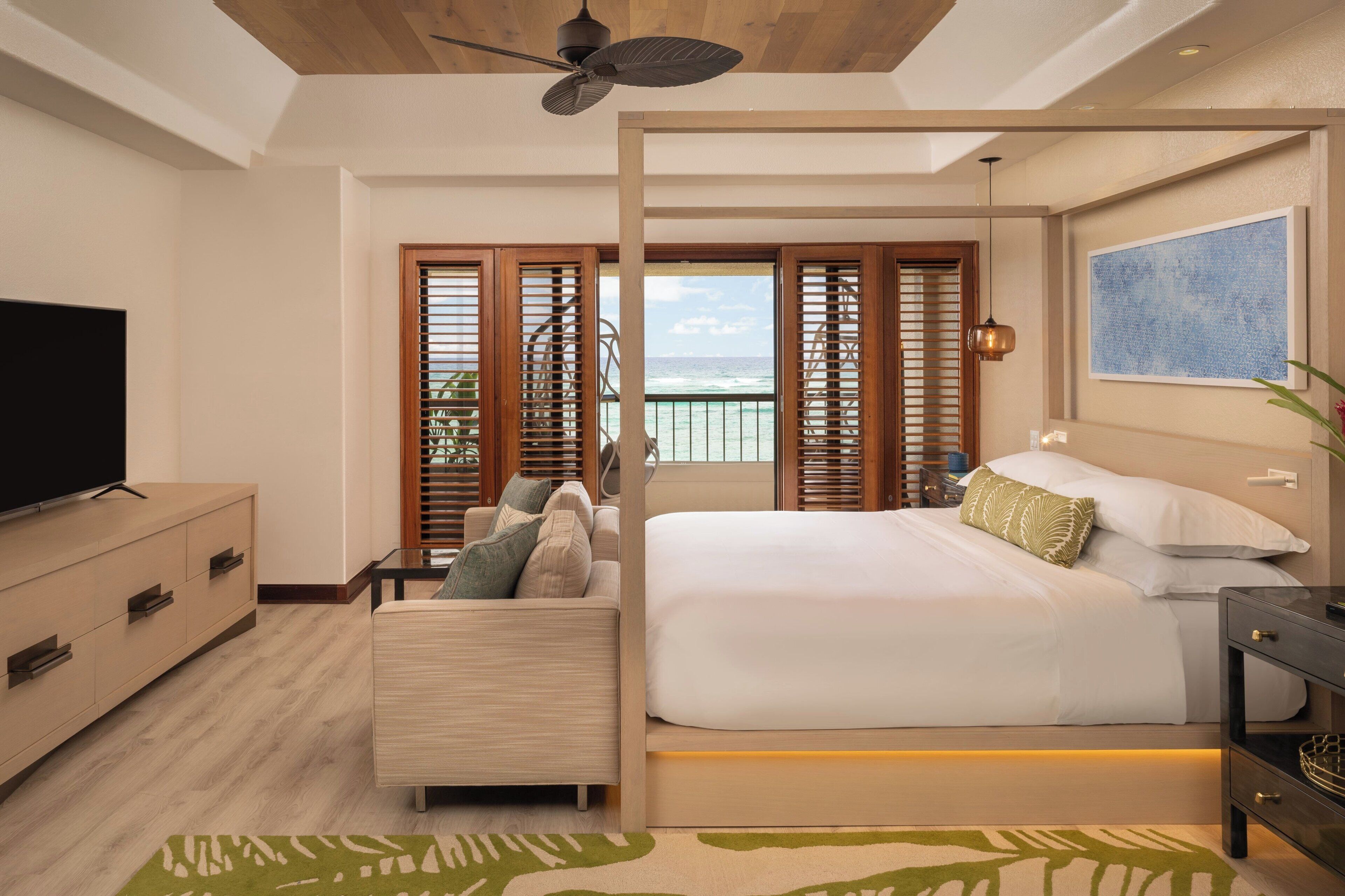 A modern hotel room with a bed, a TV, a sofa, and a balcony with ocean views.