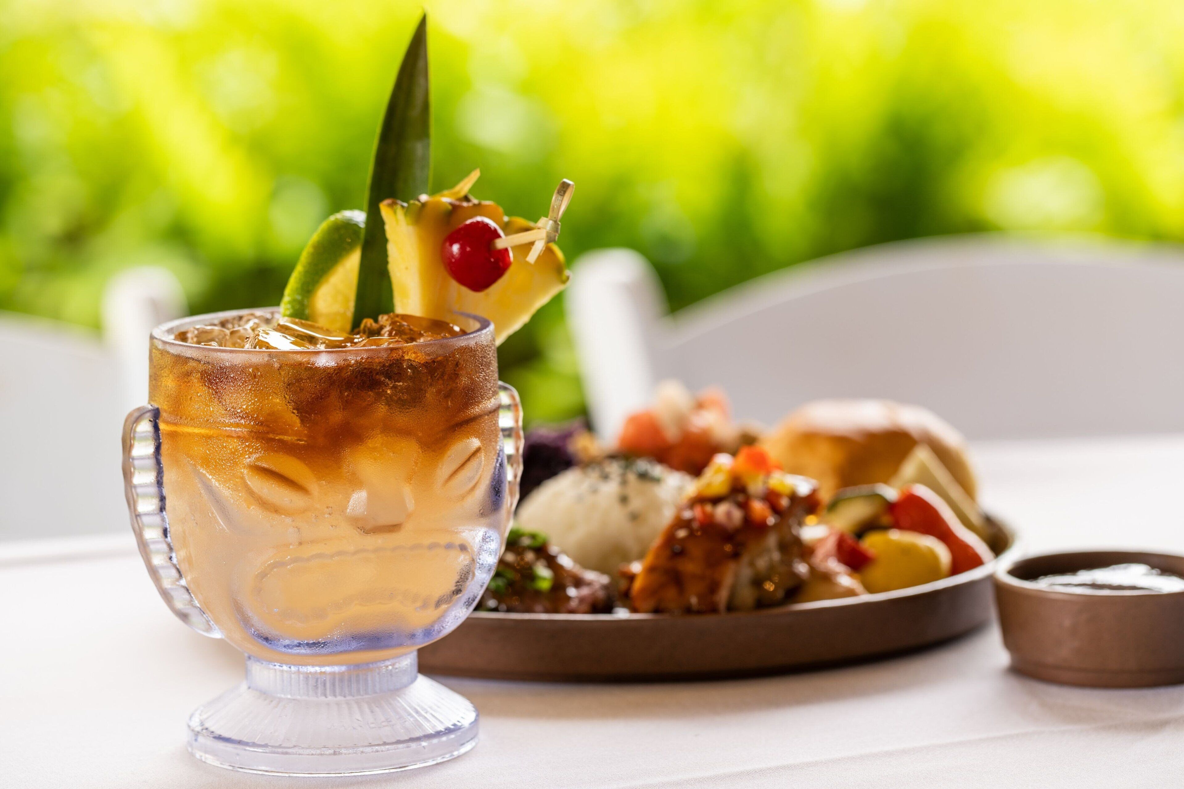 A glass of iced beverage with a lime wedge and cherry garnish, a plate of mixed vegetables, and a small bowl of sauce on a table.