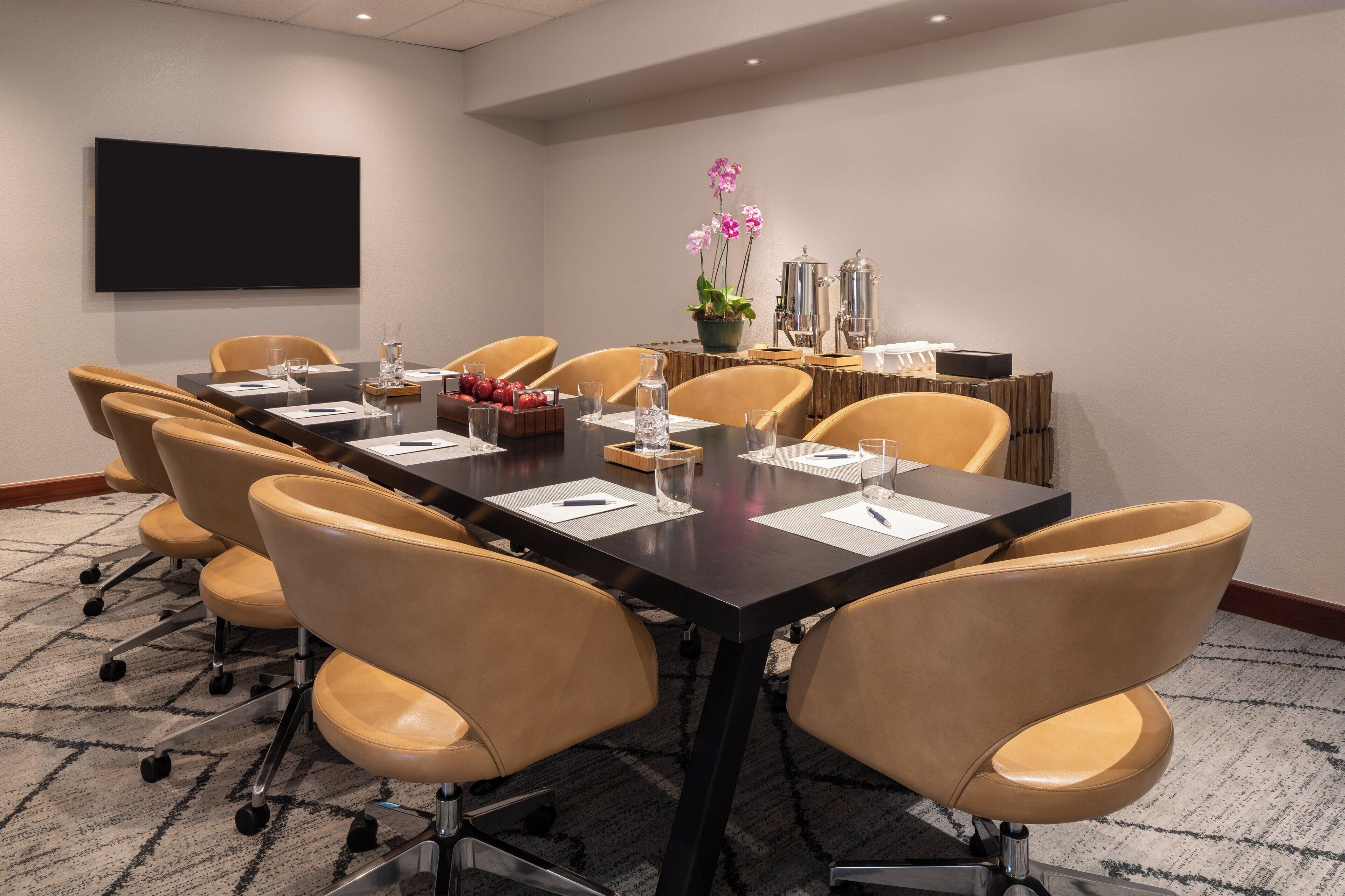 A conference room with a long table, six chairs, a flat-screen TV, a coffee station, and a decorative plant.