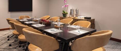 A conference room with a long table, six chairs, a flat-screen TV, a coffee station, and a decorative plant.