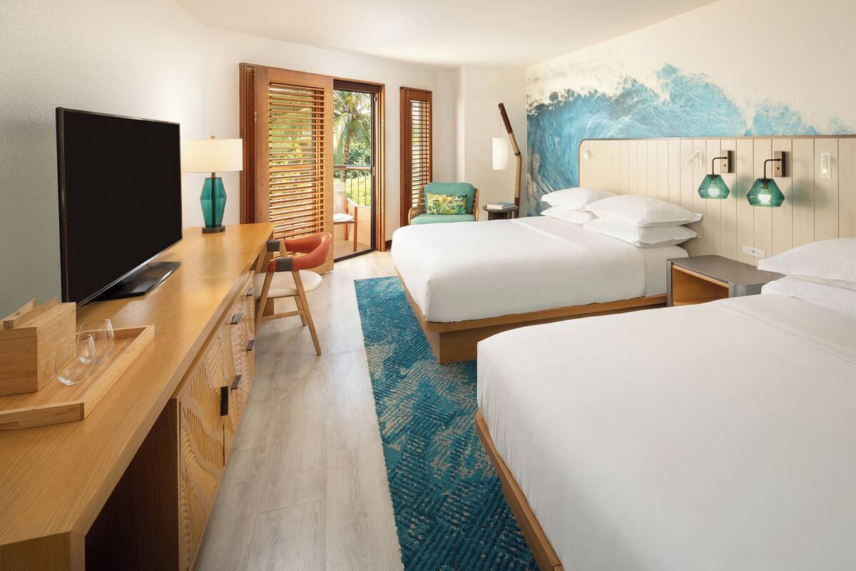 A hotel room with two beds, a TV, a desk with a laptop, and a balcony with a view of palm trees.