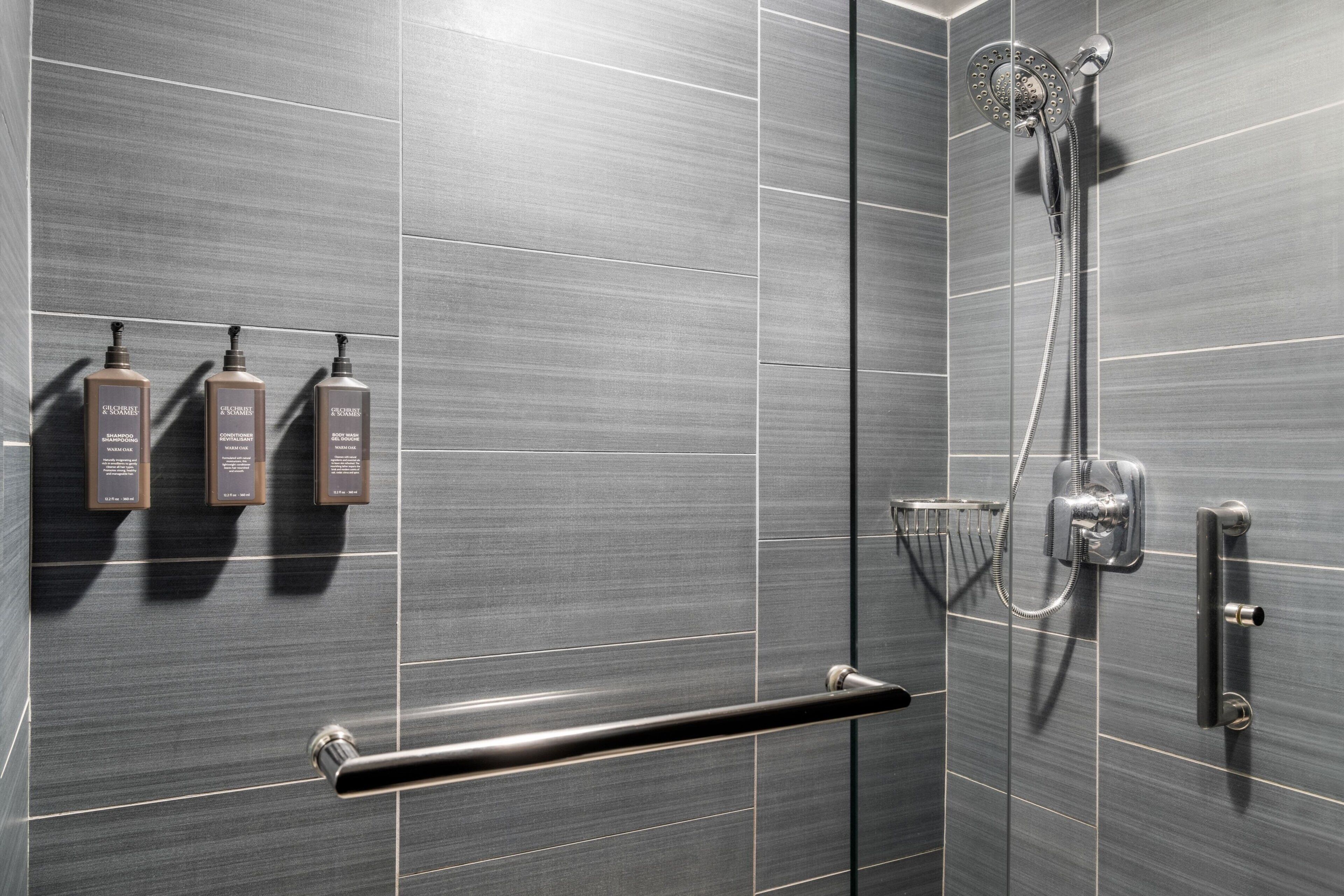 Een moderne badkamer met grijze tegels, een handdouche, een handdoekhouder en drie flessen shampoo.
